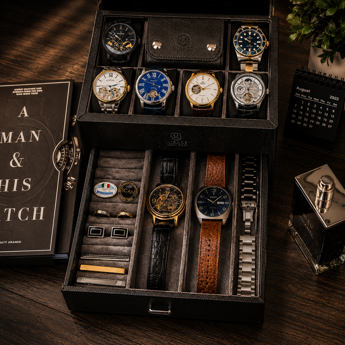 An open black watch storage box displayed on a wooden table, holding multiple wristwatches, cufflinks, and slim accessories. A watch box insert and a small calendar are placed beside it, with a perfume bottle nearby.